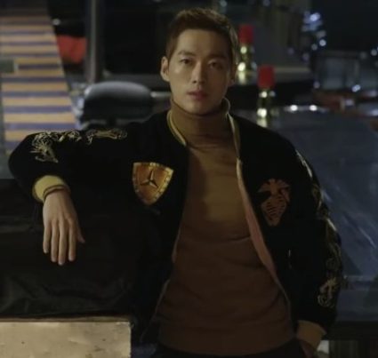 Nam Goong mean in a navy and gold varsity jacket leaning on a counter